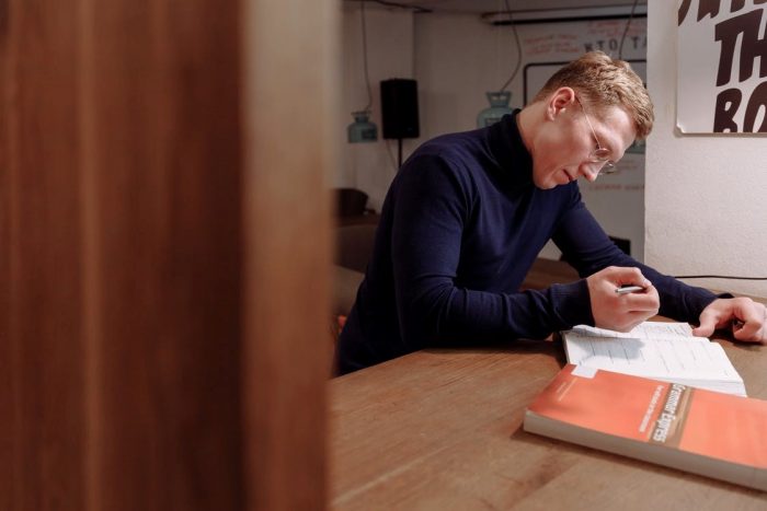 A Bespectacled Man Writing in a Book Beside a Closed Grammar Book On the Desk